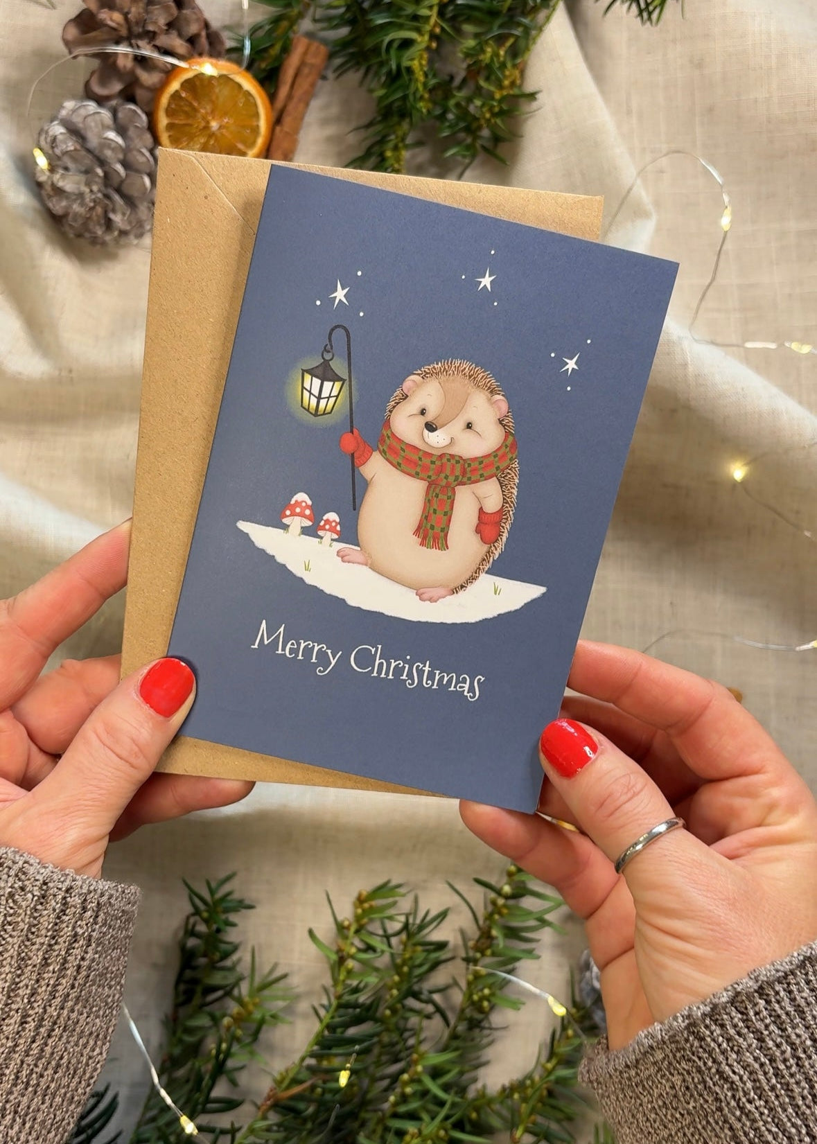 Person holding a Christmas card with an illustration of a hedgehog wearing a scarf and mittens, surrounded by festive decorations.